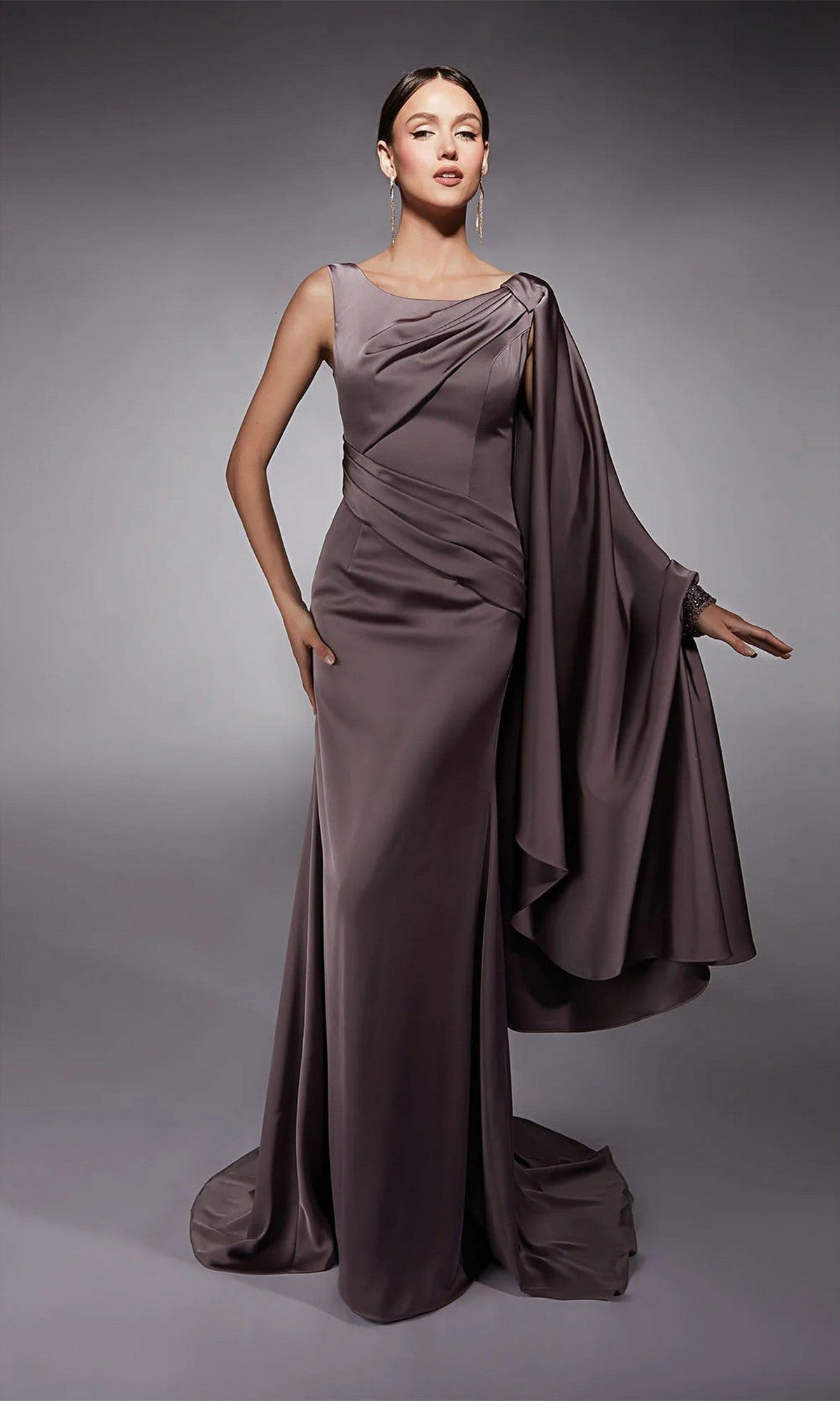 A woman wears the Alyce 27722 long formal dress in Smoke, featuring a floor-length satin design with a sleeveless cut and an asymmetrical draped cape on one shoulder, set against a plain gray background.